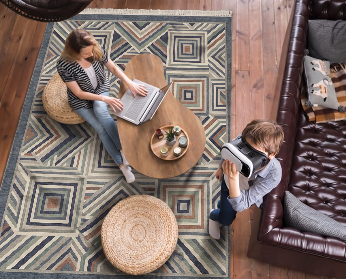 Slate-bordered hand-tufted wool rug with concentric squares and chevrons in slate blue, sage, rust and ivory, seen from above with two people working on laptops